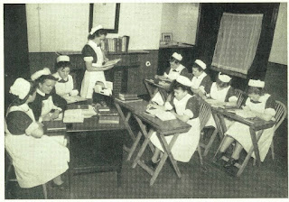 Photograph shows nurses receiving instruction at the Adelaide Hospital, Dublin, Ireland, c. 1950. Five student nurses sit at two rows of desks, facing a senior nurse seated at a larger executive desk with two other nurses at her shoulder. One desk is empty and the former occupant, a student nurse, is apparently reading something aloud to the other nurses. 