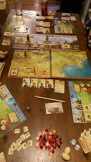 A game of Near and Far in progress. The entire table is covered with components. There are cards, plastic gems, various tokens, and many other items besides around the play area.