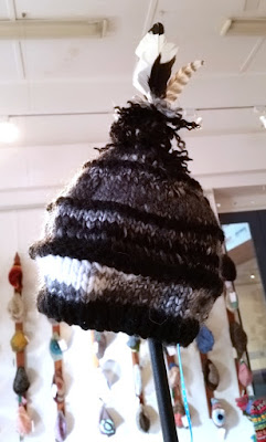 A black and white tall knitted beanie with stripes of different widths and topped with natural magpie feathers.