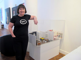 Woman standing next to a display caseWoman standing next to a display case in a gallery with two 1/12 scale miniature scenes in it" one a cafe, and one a holiday cottage. with two 1/12 scale miniature scenes in it" one a cafe, and one a holiday cottage.