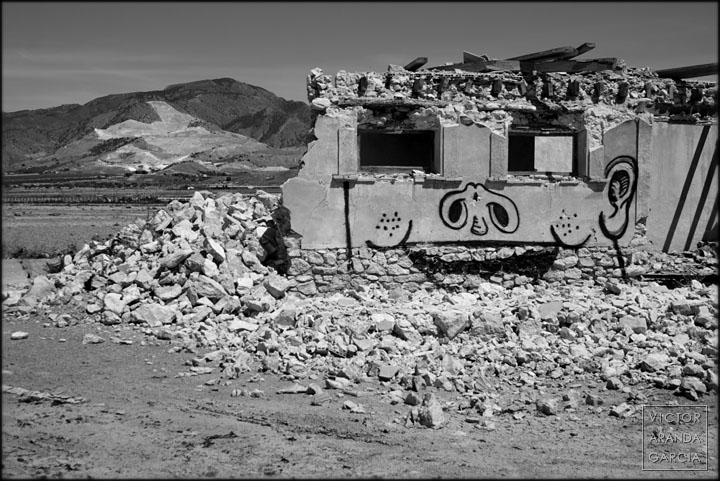 De la montaña a la casa, de la casa a la tierra: anatomía de una ruina Fotografia de una casa en ruinas con un grafiti y una montaña con una cantera al fodo