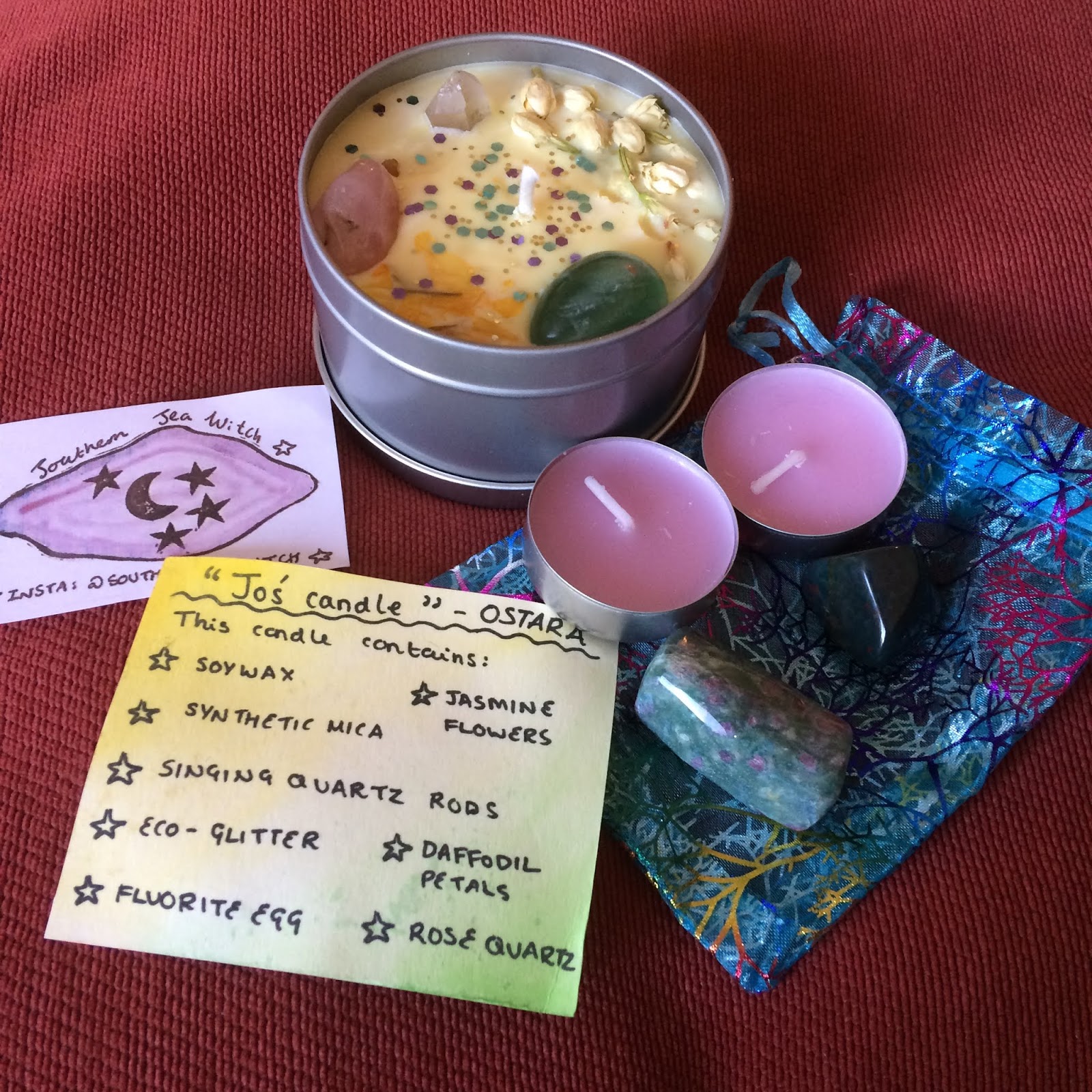 A photo featuring the open candle with it's lid off, the Southern Sea Witch logo card to it's left, the tealights and crystals on top of their pouch on the right, and the handwritten list in front