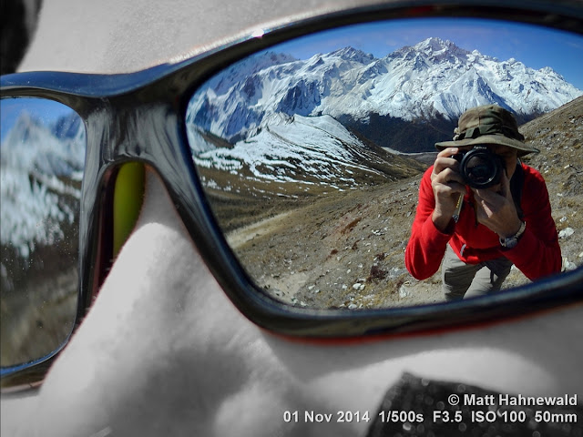focal black and white; Himalayas; mirrored sunglasses; reflection; Photoshop; edited; Langtang Trek; Kyanjin Gompa; Kyanjin Ri; mountains; background; Matt Hahnewald Photography; Facing the World; photography; photo; image; Nikon D3100; Nikkor AF-S 50mm f/1.8G; prime lens; 50 mm; 4 : 3 aspect ratio; horizontal format; closeup; portrait; portraiture; street portrait; sunglasses; trekking; outdoor; colour; colourful; character; real people; human; human head; male; holding Nikon D3100 DSLR camera; consent; rapport; encounter; environmental portrait; phototourism; travel; travel portrait; travel destination; international; Nepal; Asia; street photographer; old man; one person; adult; posing; incredible; determined; focused; fun; friendly; male street photographer; photographing; snapping; shooting; taking a photo; both hands; amateur; sunshine; snow