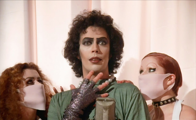 Left to right Patricia Quinn, Tim Curry, and Nell Campbell from The Rocky Horror Picture Show (1975). Tim Curry is wearing make-up and pearls and is dressed in operating room scrubs. He holds his right arm, covered in a sleeve of glittery fabric, up to his chin and stares out at the audience with a look of hopeful anticipation. Patricia Quinn and Nell Campbell are both looking up at him with their hands on his shoulders and wearing pink rubber surgical masks over their faces.
