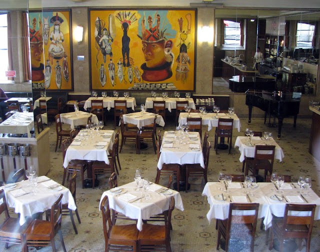 Salão de restaurante com mesas e cadeiras, piano e grandes quadros de índios na parede