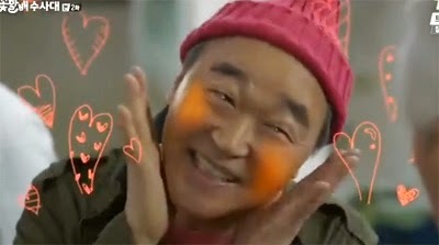 Kang Suk strikes an aegyo pose while pink hearts spring up around him.