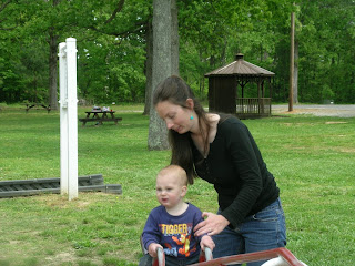 Image shows a woman standing in a park with a toddler. In the background are several picnic tables