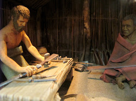 A full-sized diorama of a pakeha man and a Maori man in a Maori hut. The pakeha is working on agun.
