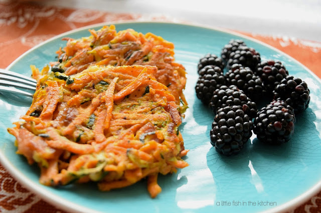 Two eye-catching sweet potato and zucchini cakes are served along side a portion of vibrant ripe blackberries an a teal ceramic plate. The sweet potato cakes are delightly colorful: shredded amber sweet potato is mingled with bits of green from the zucchini mixed in perfectly formed round, veggie pancakes.
