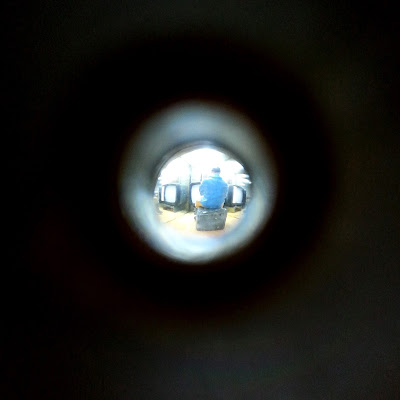 Tiny miniature man in coveralls, sitting in front of a bank of miniature TV sets, seen through a peep hole.