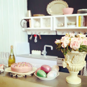 Modern dolls house miniature kitchen in colours of white, grey, black and pink. In the foreground is a vase of roses and an easter afternoon tea laid out.
