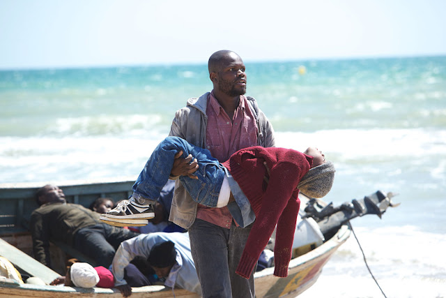 Vor einem schlichten Holzboot, in dem leblose Körper liegen, steht ein Mann, der einen kleinen Jungen in den Armen trägt. Hinter ihnen das offene, türkisfarbene Meer ...