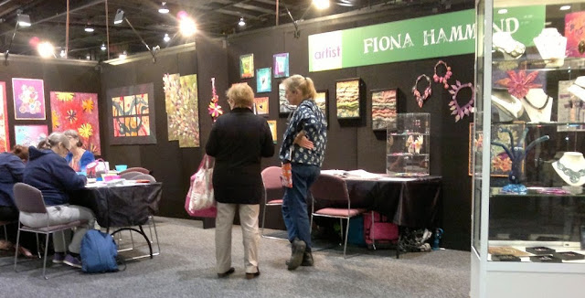 Fiona Hammond's stall has mixed media wall art and necklaces displayed on the walls with sculptural pieces and jewellery in display cabinets. A large display cabinet features on the right hand side. On the left of the photograph are a table and chairs where Fiona can be seen wearing a royal blue top conducting a beading class.  Her sister Kate is standing in the centre wearing a patterned top and her hair in a blonde ponytail, talking to a visitor. A large green sign acros the top of the stand has "Fiona Hammond" emblazoned on it.