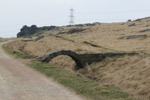 A track running around the gristone outcrop that forms the edge. A small, arched bridge crosses a stream running alongside the path.
