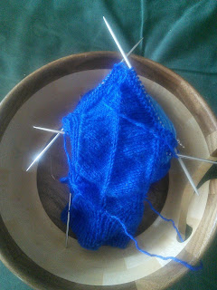 A sock in blue yarn on double-pointed needles.  The cuff of the leg is done, and there are a few rows of garter stitch short-rows at the beginning of the heel.   The sock, along with the ball of working yarn, is in a wooden yarn bowl.