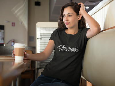 Survivor tee - Audacity Tees Woman sitting in booth with coffee wearing black survivor tshirt