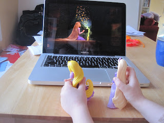 Two Rapunzel dolls being held in front of a laptop showing Rapunzel from Tangled