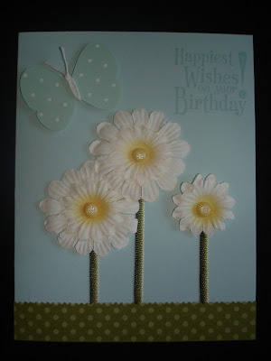 Light blue card with patterned green paper as grass, a punched butterfly and three paper daisies with green ribbon stems. 