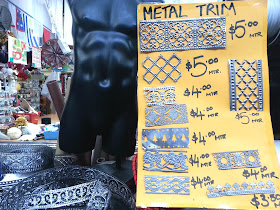 Poster displaying various metal trim pieces, with prices per metre, with lengths of the trims coiled on the table beside it.