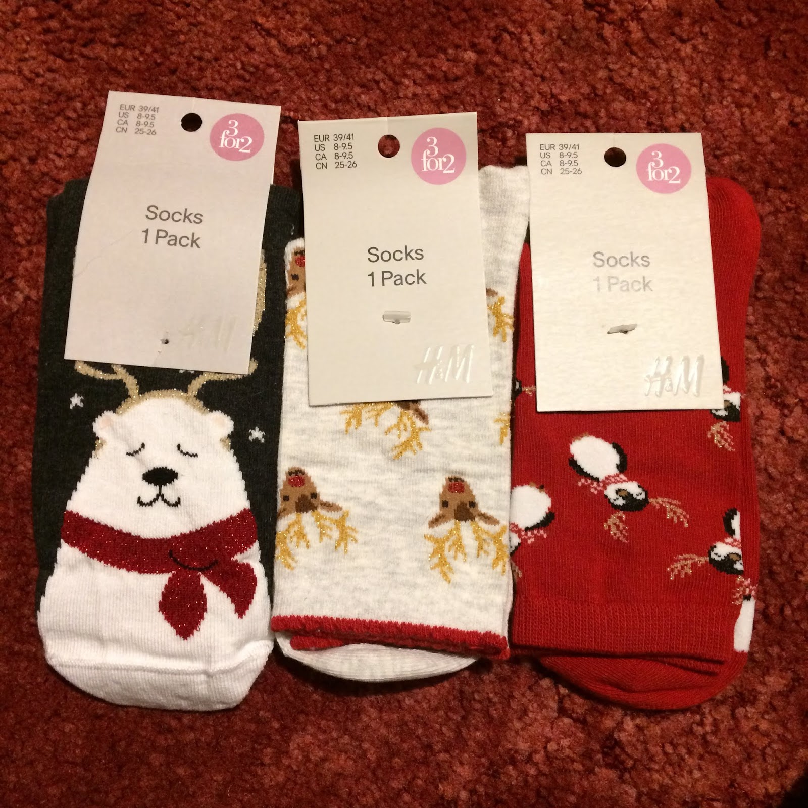 Three pairs of festive socks, one navy blue pair featuring a polar bear weaing a red scarf and antlers, a pale grey pair with a repeated pattern of Rudolph's head, and a bright red pair with a repeated pattern of a penguin wearing antlers.