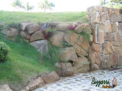 Detalhe do acabamento do canto do muro de pedra faceado e no início do talude o muro de pedra sem cimento com nichos para colocação de plantas com o piso de pedra bruta.