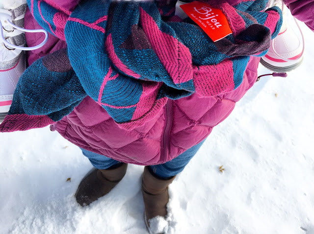 [Image looking down at the pink and blue messy double knot of a rucksack tied in front over a pinkish-purple coat. The carrier happens to match the wearer’s cost and blue jeggings. Orange-ish red tag with white text BijouWear visible on top of knot. Toddler feet in white boots are at Mama’s sides. Farther down are Mama’s brown boots in the freshly fallen white snow.]