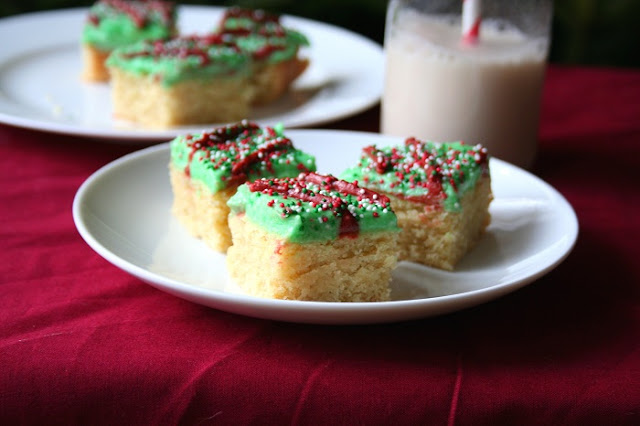 Sugar Cookie Bars – Low Carb and #glutenfree from All Day I Dream About Food inkatrinaskitchen.com #BringtheCOOKIES low carb and gluten free sugar cookie bars with green frosting and nonpareil sprinkles on a white plate and red tablecloth and milk in the background