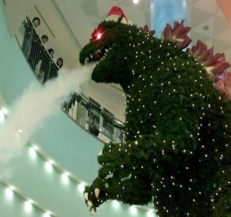 A giant tree with little white ornaments shaped like Godzilla and shooting smoke from its mouth. It's in a mall and people are looking down on it from a railing.