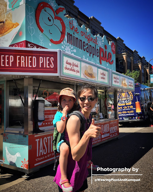 [Image of me, a light tan skin Asian woman with sunglasses and a smile, giving the viewer a thumbs up while wearing a preschooler on my back in a green linen onbuhimo. We're standing in front of a large iconic food truck that says The Original Minneapple Pie.]