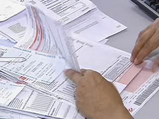 A imagem mostra alguém organizando as contas para serem quitadas, mas ainda não recebeu o salário do mês. Olhar para a gaveta das correspondências, e está cheia de todos os boletos a serem quitados no mês, mas a sua conta bancária está além do vermelho; em razão dos atrasos e parcelamentos do salário.