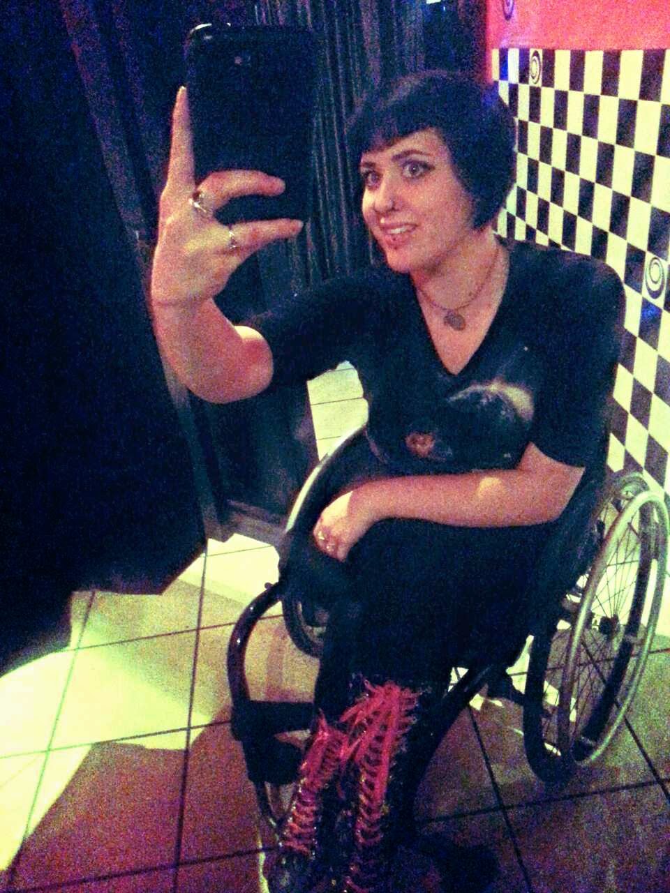 Woman with short black hair, wearing dark patterned clothes and tall boots with red laces, sitting in a a wheelchair, with hand up holding iPhone