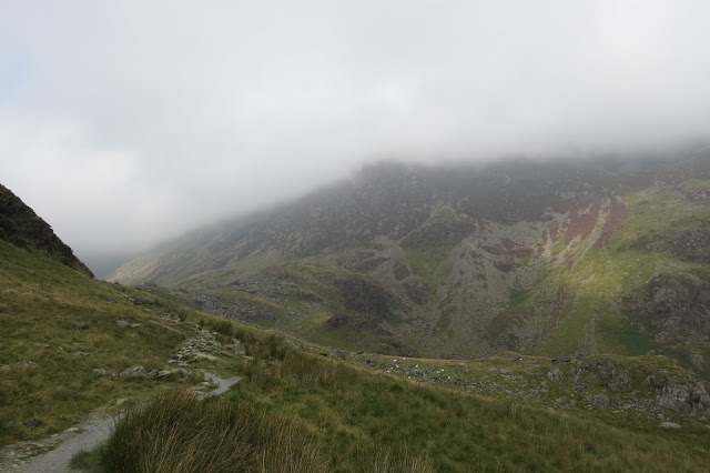 A path leads round a bend towards a valley halfway up the mountain. The cliffs are wreathed in cloud.