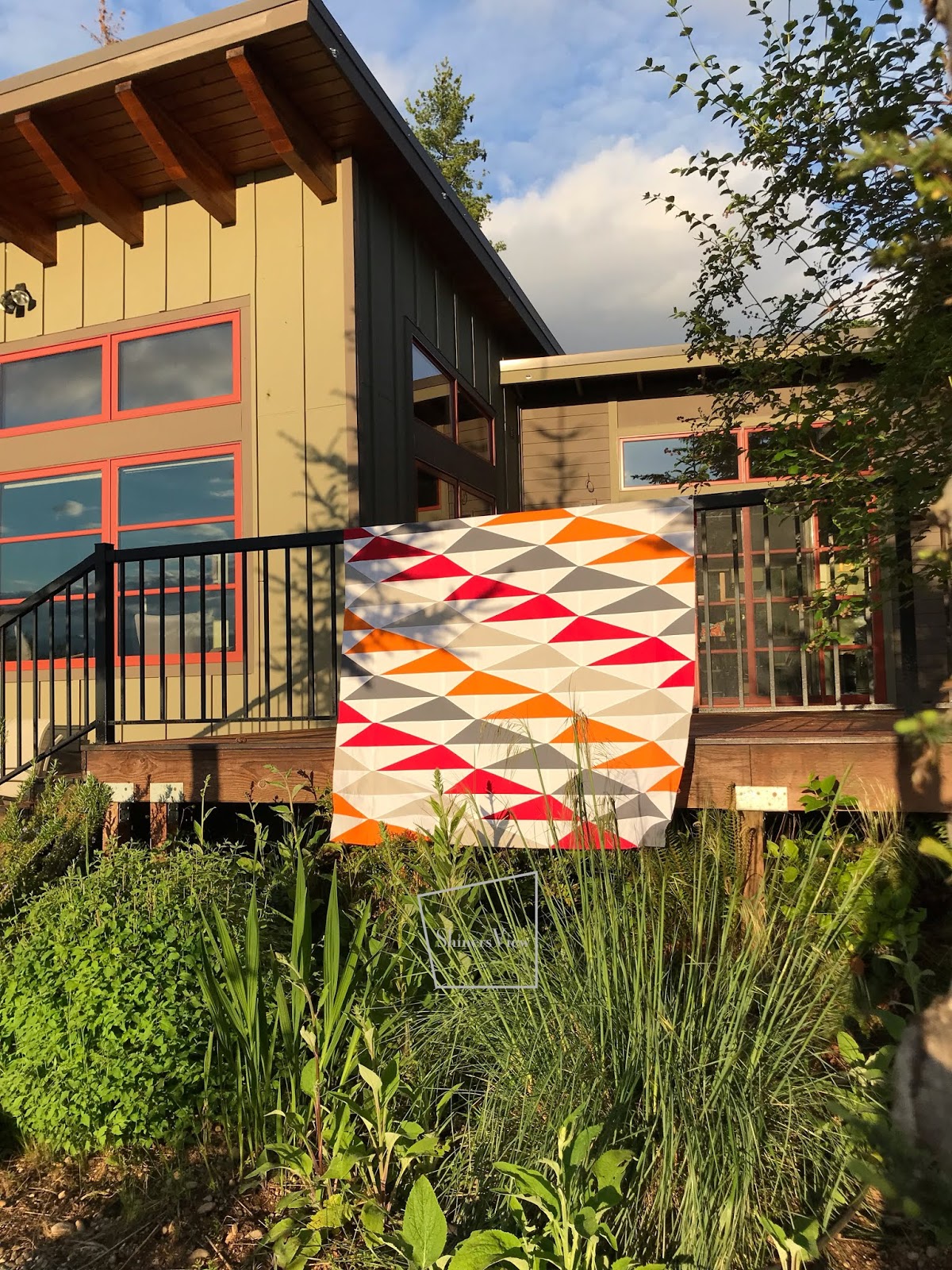 photo of a orange and red quilt top laying on a deck railing with a green modern house in the background