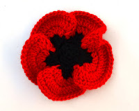 Crocheted poppy. Black centre has 5 blunt points around which are rounded red petals which overlap each other and curl upwards.
