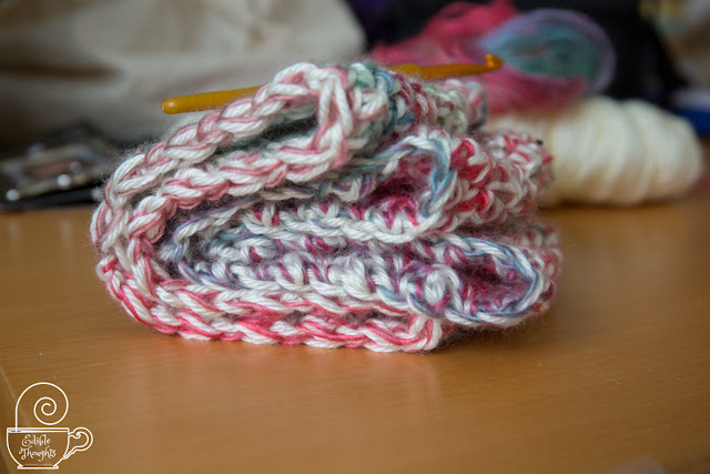 Image of two toddler-sized multi-colored cowls folded over on each other on a wood table with a golden crochet hook on top and the skeins of yarn used to make the cowls out of focus in the background.
