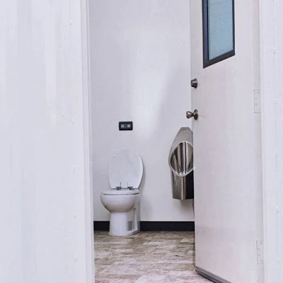 Foto de um vaso sanitário comum, dentro de um banheiro. Há um mictório de alumínio ao lado do vaso.