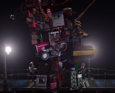 Janet and Michael are standing on a metal bridge-type thing that has some street lights on it. They're separated from the Judge by a giant pile of stuff, including an oven, a giraffe, and a tractor. 