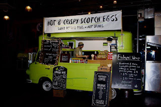 A large white rectangular van with a white rectangular sign with hot and crispy scotch eggs in black casual font with a large pale man inside wearing a wjite and red hat and white shirt and black jeans on a bright background 