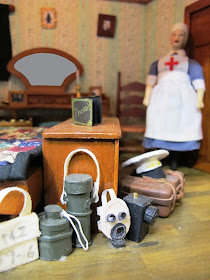 One-twelfth scale miniature bedroom scene with a nurse in the background, and a pile of luggage including a gas mask and military items in the front.