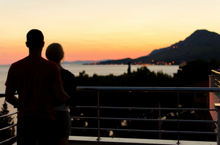 A couple stand on a balcony and look out at the sunset.