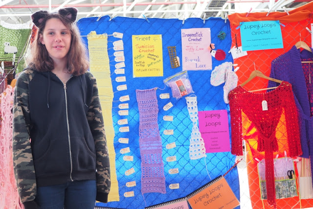 Miss E. is standing on the left hand side. She is wearing fluffy 'cat ears' on her head, a black jacket with camouflage patterned sleeves and blue jeans. She is standing in front of the main display which has samples of stitches, garments, tools and some display books pinned and hung. The yellow sign says "Tricot, Tunisian crochet, afghan stitch, shepherd's knitting, Scottish knitting, idiot stitch, railway knitting". The pink sign says "Broomstick crochet, jiffy lace, peacock lace" The pink/purple and blue signs each say "Lupey Loops, www.lupeyloops.blogspot.com.au, crochet, tricot, hairpin, broomstick". 