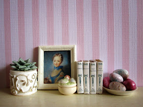 Modern miniature dolls' house scene with a white shelf in front of pink striped wallpaper. On the shelf is a succulent in a white and gold pot, a blue painting of a lady, a set of Jane Austen novels and a bowl of pink and white Easter eggs,