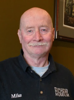 Man with a mustache wearing blue shirt with the words "Pennsylvania Military Museum" and his name "Mike" embroidered on it