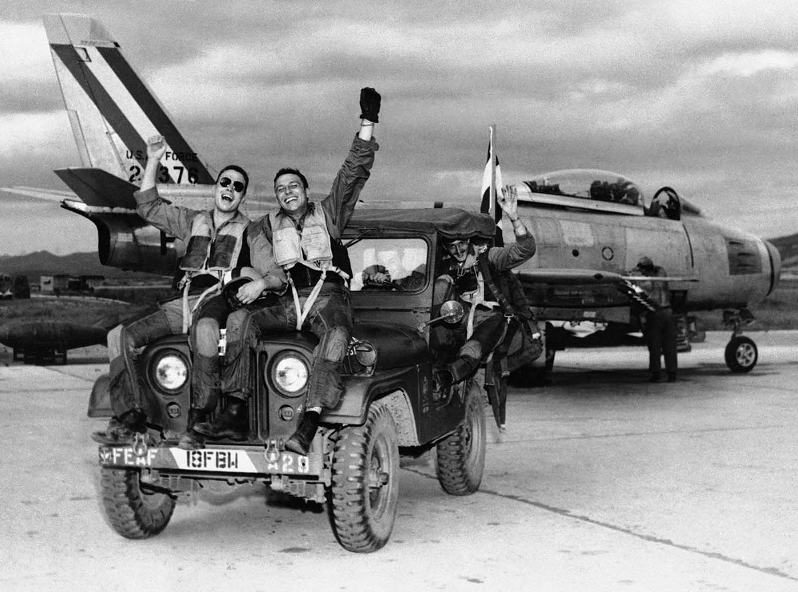 Three happy fliers of the 18th Fighter Bomber wing let the world know how they feels as they returned from a combat mission over North Korea to learn of the armistice signing on July 29, 1953. Left to right are: 2nd Lt. John Putty, Dallas, Tex.; 1st Lt. James A. Boucek, Ottawa, Kansas,: and 1st Lt. Richard D. Westcott, Houston, Tex., waving from the back seat of the jeep.