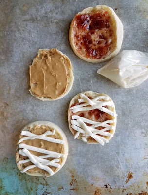 These super simple English muffin mummies are a perfect way to start Halloween. Just a couple of minutes and a few ingredients is all it takes to get the day off on an undead foot! baggie of cream cheese mixture piping out bandages over english muffins