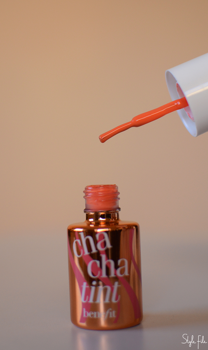 An image of Benefit cha cha tint in a shiny glass bottle with a white cap and brush in front of a peach background for a beauty review