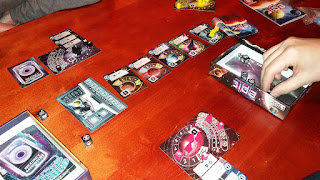 A game of Tiny Epic Galaxies in progress. Each player has their play mat, decorated with a galaxy surrounded by a track used to indicate, by means of wooden tokens, how much power and how much culture that player has. Four more tracks curve along the right side of the mat to indicate how advanced the civilization is, with corresponding increases in fleet size, dice to roll, and victory points. Sticking out from under the player mat are cards representing colonised worlds. Five more cards are arranged in the centre of the table to reperesent planets available for colonising. The players have rocket ship tokens, some on their player mats, some on the available cards in the centre, that they move in the course of the game.