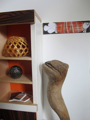 Close up of a modern dolls' house miniature scene of a 1970s display cabinet holding a selection of art objects and books. Next to it is a wooden sculpture, and an aboriginal artwork on the wall.