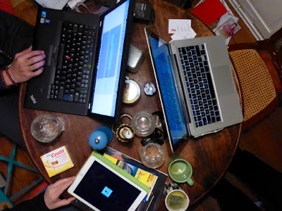 Hände, zwei Computer, ein Tablet, Wassergläser, Teebecher, Pfeffermühle, Salz, Kerze, Vitamine, Visitenkarten, Stühle (angeschnitten), Kataloge auf Bank und Boden