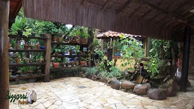 Detalhe da execução do paisagismo com as pranchas de madeira com os vasos de barro, os canteiros de pedra com plantas, o laguinho de carpas com pedra do rio com a bica d'água no pote de barro e o piso de pedra caco de São Tomé.